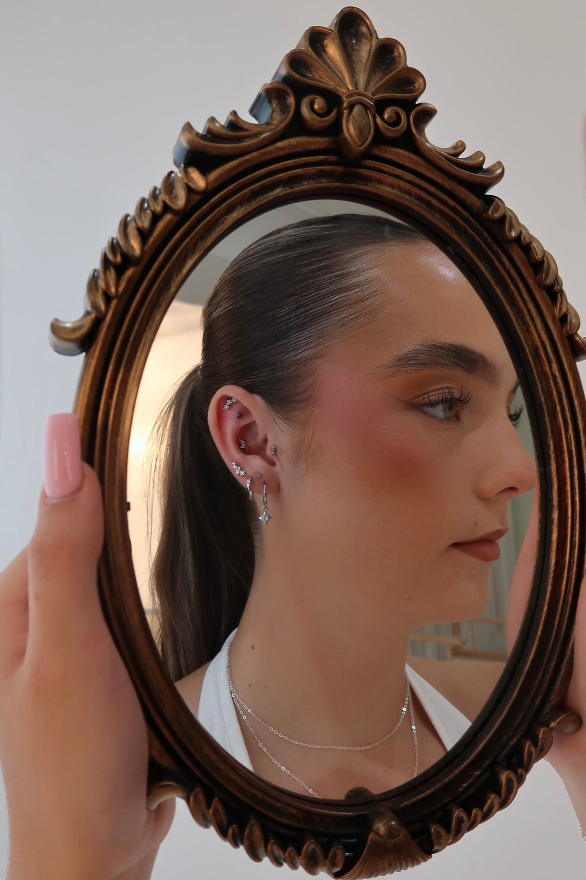 A person holds an ornate oval mirror with a dark carved wooden frame, showing their reflection. The person has dark hair in a ponytail, pink blush on their cheeks, and is wearing silver hoop earrings and layered necklaces.