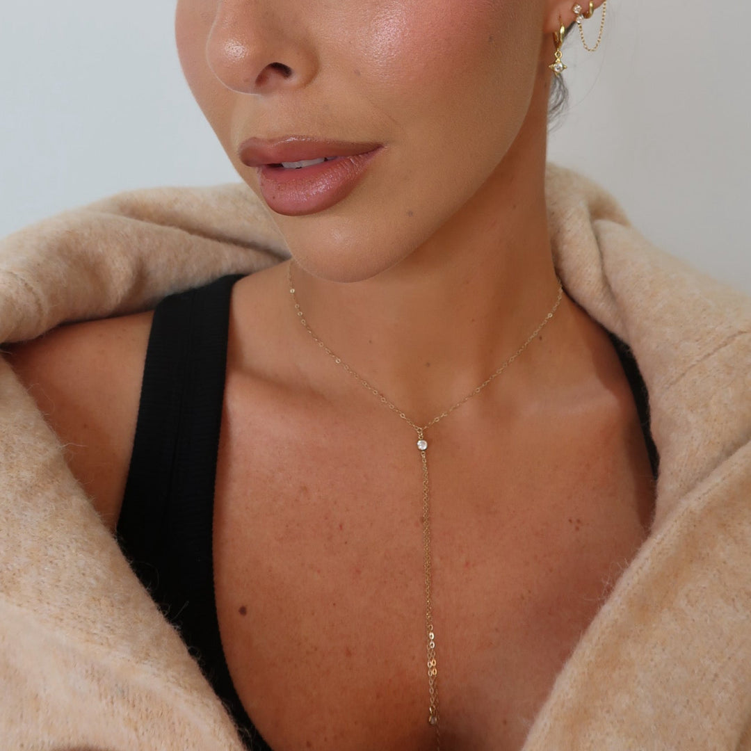 A close-up of a woman wearing a delicate gold Y-necklace with a small clear stone pendant, styled with a black top and a beige knit cardigan.