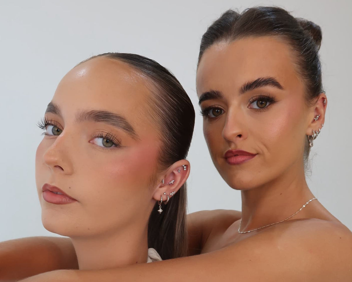 Two women posing together in white strapless dresses against a plain light gray studio background.
