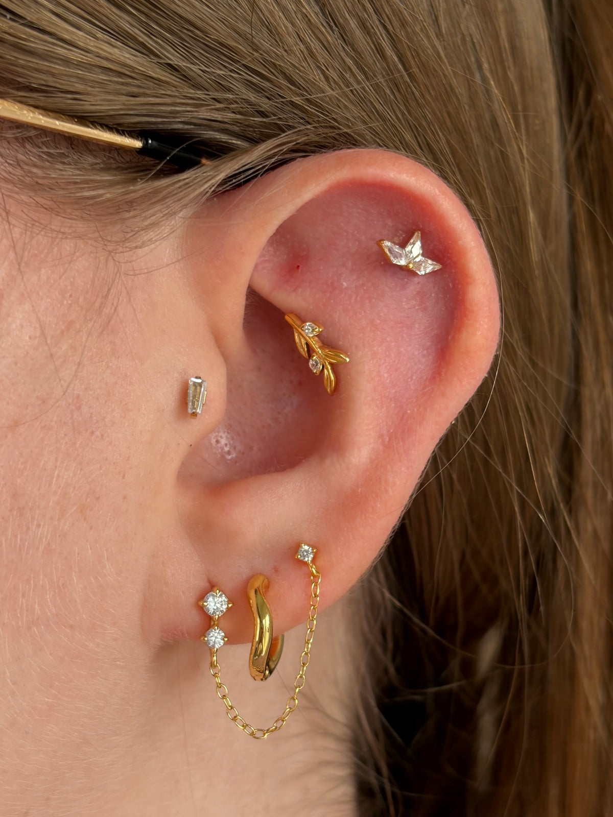 Close-up of a person's ear showcasing multiple gold and silver cartilage piercings, including a leaf-shaped cartilage hoop, a small silver stud, a gold huggie hoop, and a gold chain-link ear cuff.