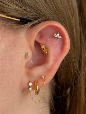 Close-up of a person's ear showcasing multiple gold and silver cartilage piercings, including a leaf-shaped cartilage hoop, a small silver stud, a gold huggie hoop, and a gold chain-link ear cuff.