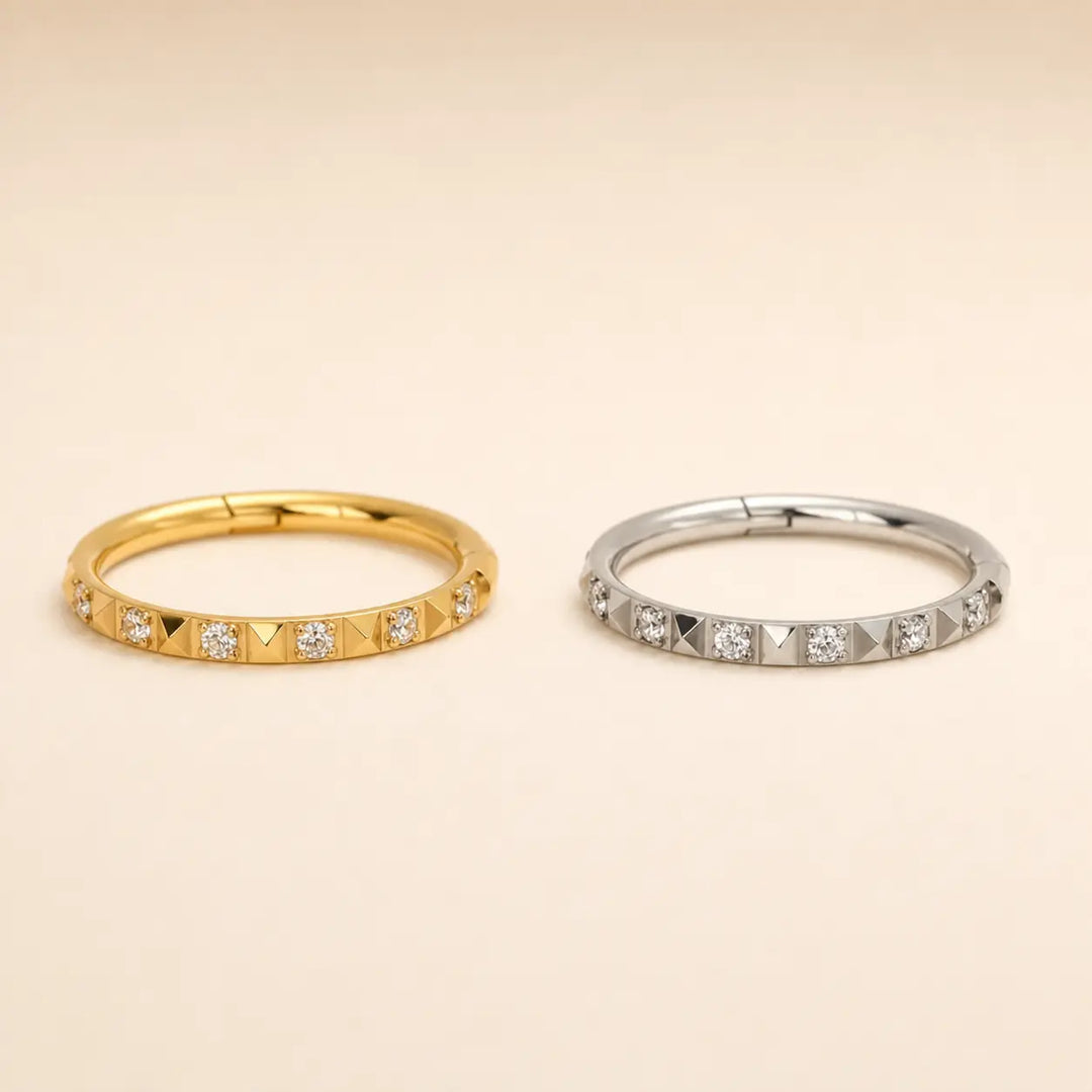 Two slim eternity-style rings on a warm beige background, one in yellow gold with small clear stones and pyramid-cut accents, and one in silver with matching clear stones and pyramid-cut accents.