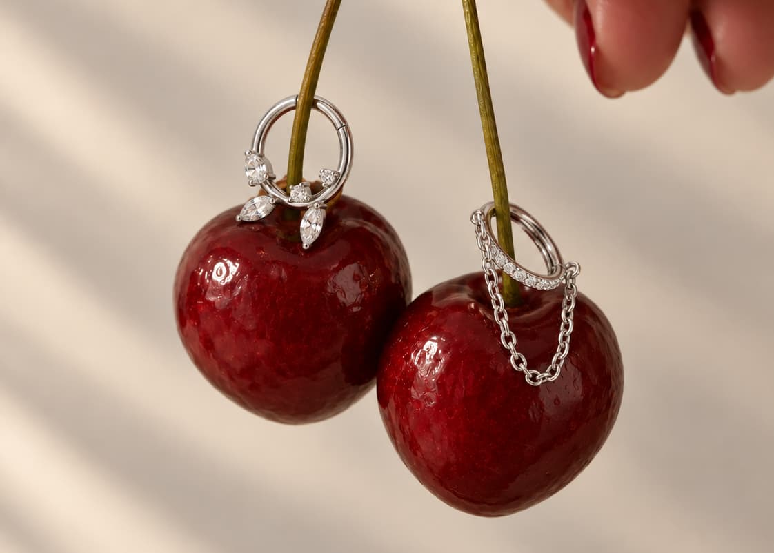 Two cherry-shaped earrings with silver rings and clear stones, hanging from stems against a softly lit beige background. A hand with red nail polish is partially visible at the top right.