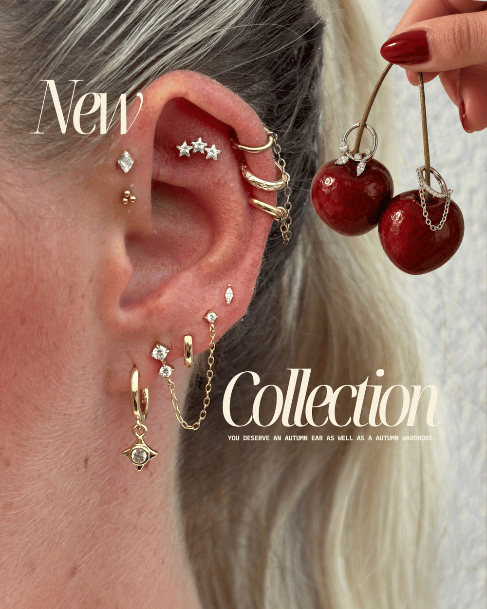A close-up of a person's ear adorned with multiple gold ear piercings and earrings, including a star-shaped stud, hoop, chain, and dangling charms. A hand with red nail polish holds a pair of dangling red cherry earrings with clear crystal accents.