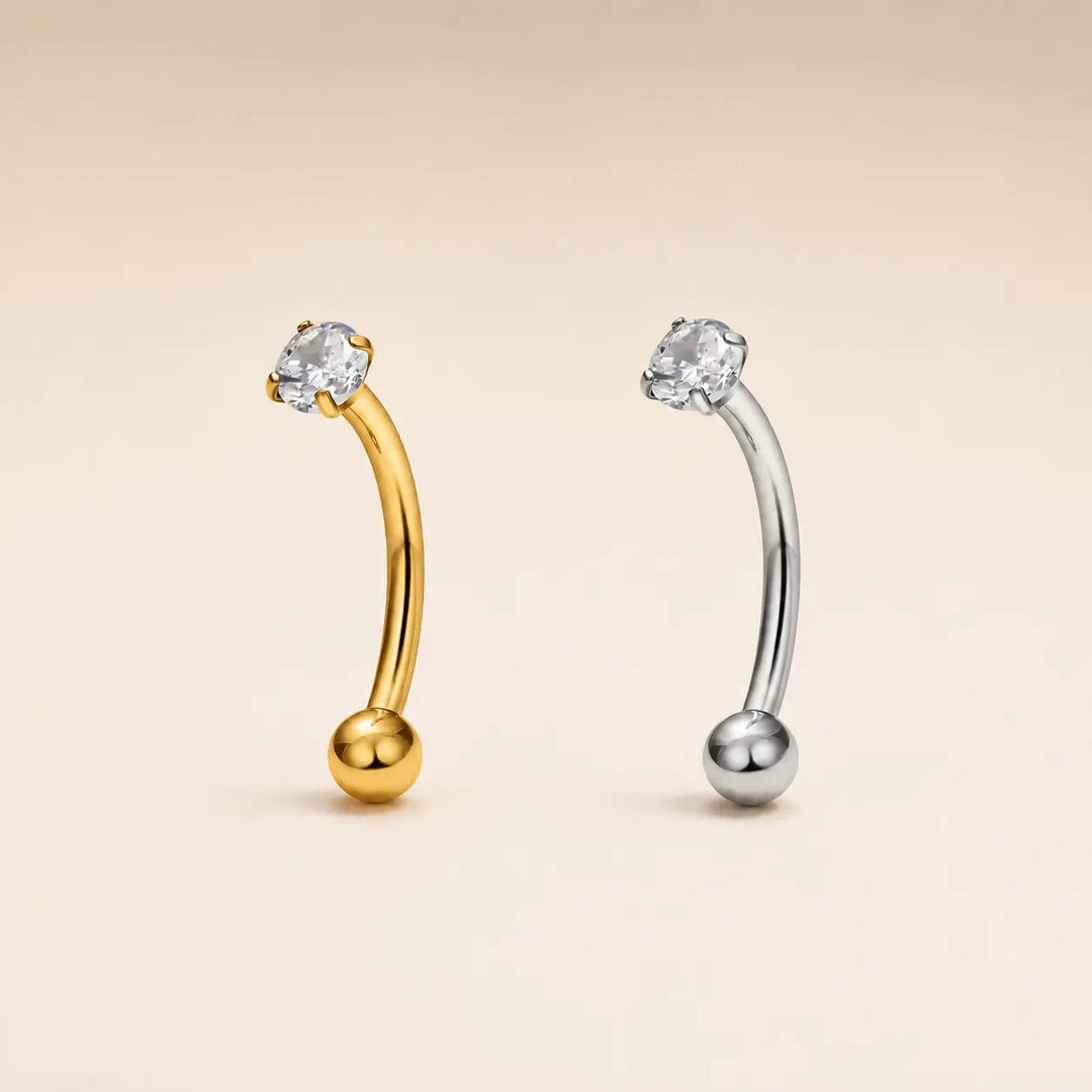 Two curved barbell-style body jewelry pieces with ball ends and a clear gemstone at the top, shown on a plain warm beige background. One is gold-toned and one is silver-toned.