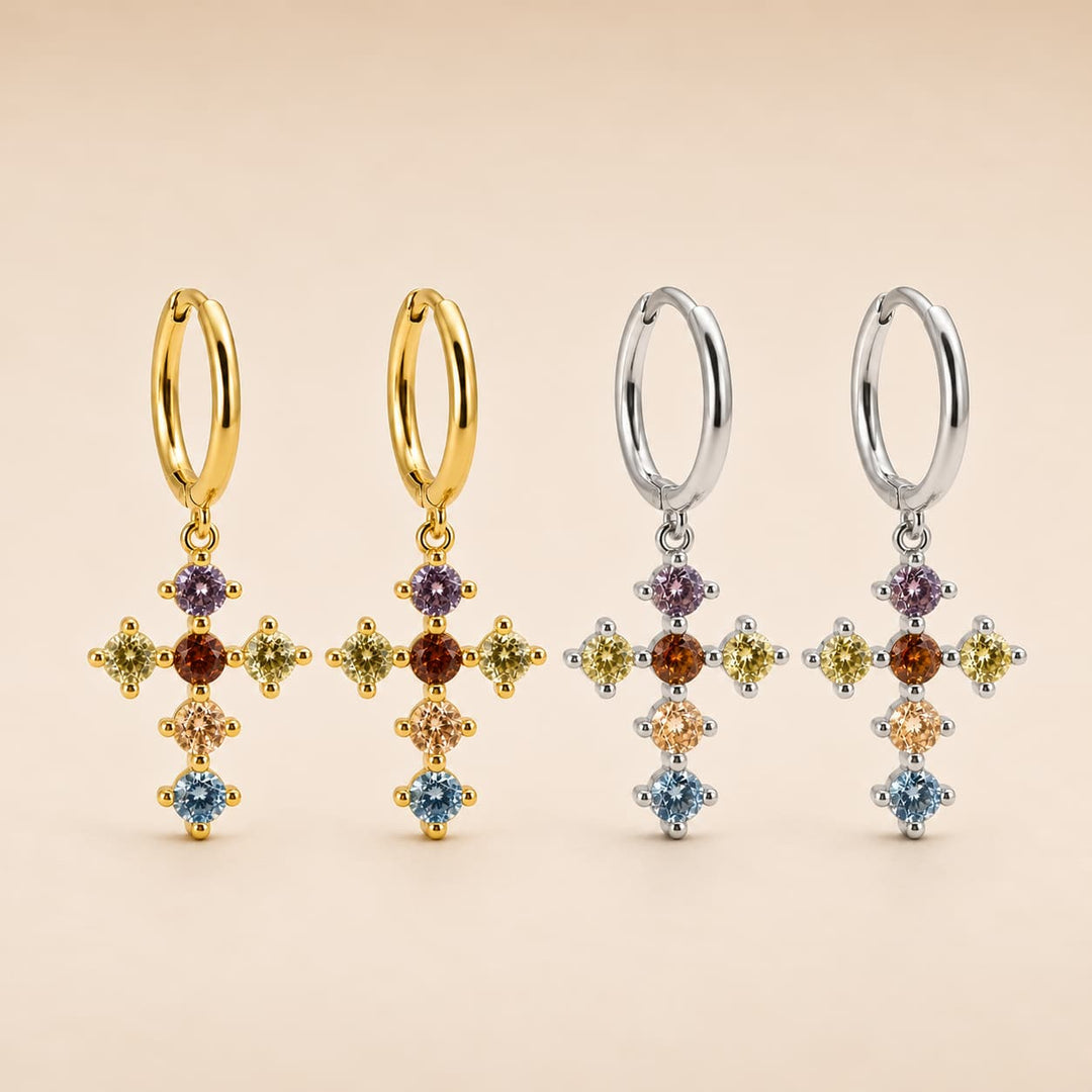 Four dangle earrings displayed on a warm beige background, featuring gold and silver hoop earrings with dangling cross-shaped charms set with multiple colorful gemstones.