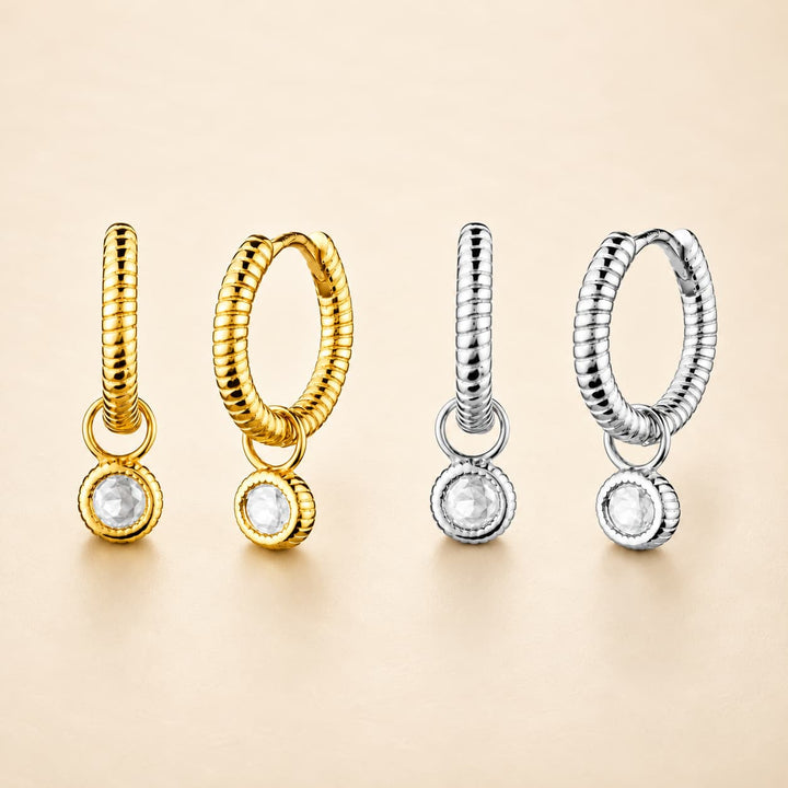 Four small hoop earrings with twisted rope-like bands and round clear stone pendants, shown in gold and silver finishes on a warm beige background.