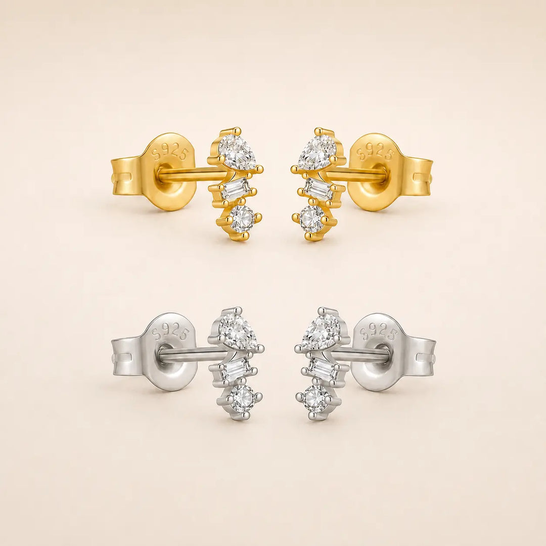 Two pairs of stud earrings on a warm beige background, one in yellow gold and one in silver, both featuring a cluster of clear baguette and round-cut stones.
