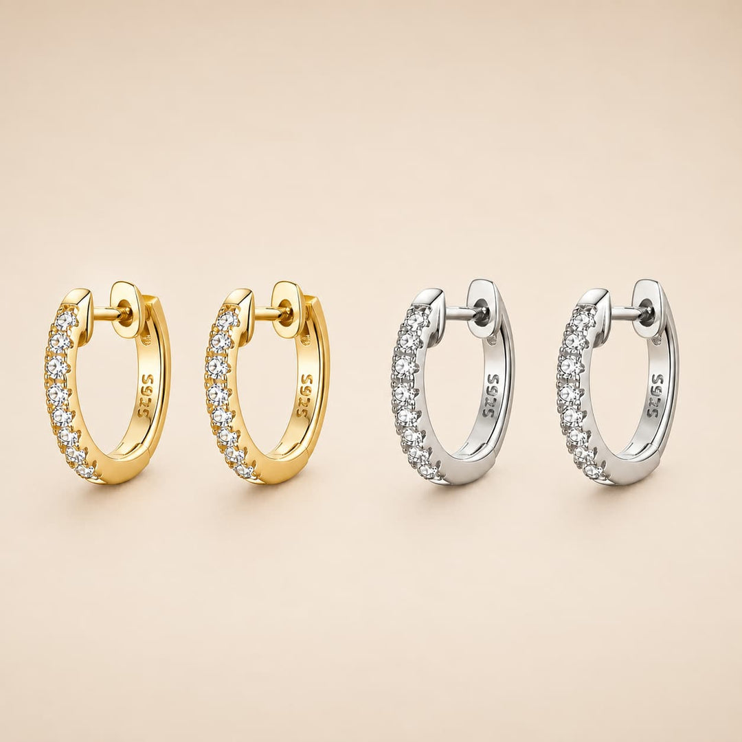 Four 925 sterling silver huggie hoop earrings, two in yellow gold and two in silver, each adorned with a row of clear pavé-set cubic zirconia stones, arranged on a warm beige background.