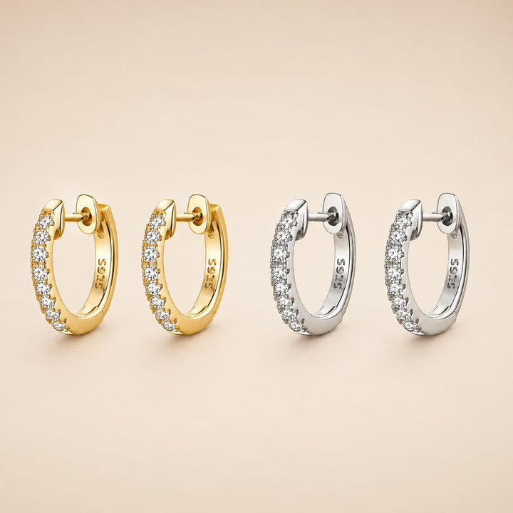 Four 925 sterling silver huggie hoop earrings, two in yellow gold and two in silver, each adorned with a row of clear pavé-set cubic zirconia stones, arranged on a warm beige background.