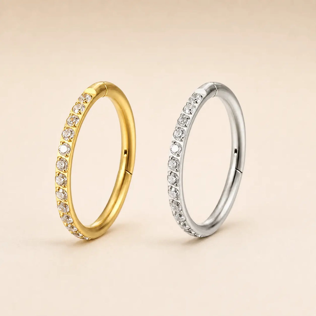 Two oval hoop earrings with rows of clear stones, shown side by side on a warm beige background. One earring is yellow gold, and the other is silver-toned.