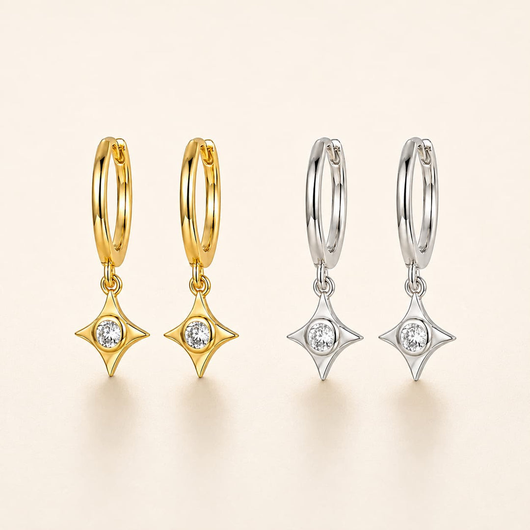 Four small hoop earrings with dangling star-shaped settings are shown on a warm off-white background. Two hoops are yellow gold and two are silver, each with a clear round stone in the center of the dangling star.