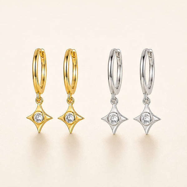 Four small hoop earrings with dangling star-shaped settings are shown on a warm off-white background. Two hoops are yellow gold and two are silver, each with a clear round stone in the center of the dangling star.