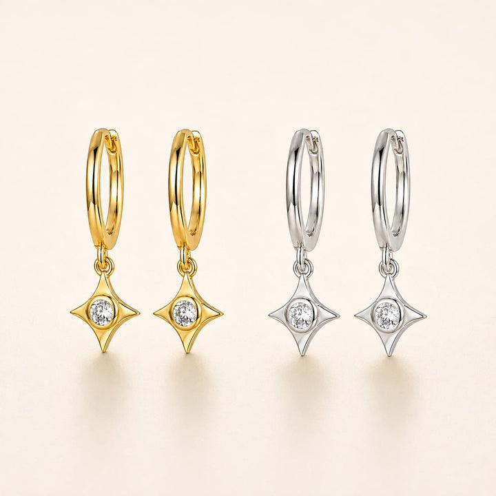 Four small hoop earrings with dangling star-shaped settings are shown on a warm off-white background. Two hoops are yellow gold and two are silver, each with a clear round stone in the center of the dangling star.