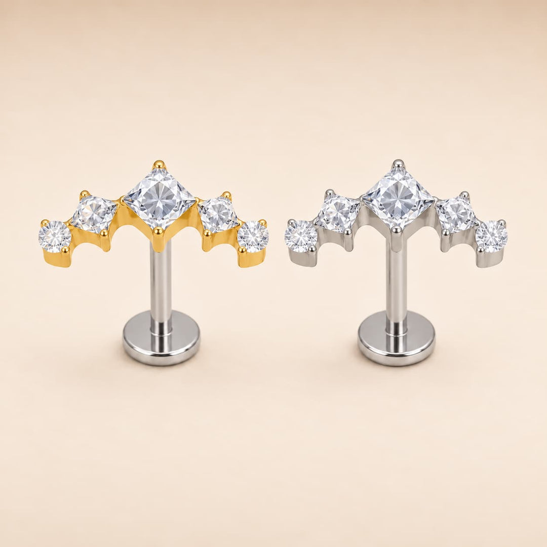 A pair of dome-shaped cartilage piercings featuring a central clear gemstone surrounded by smaller clear stones, with one earring in yellow gold and the other in silver, displayed on a warm beige background.