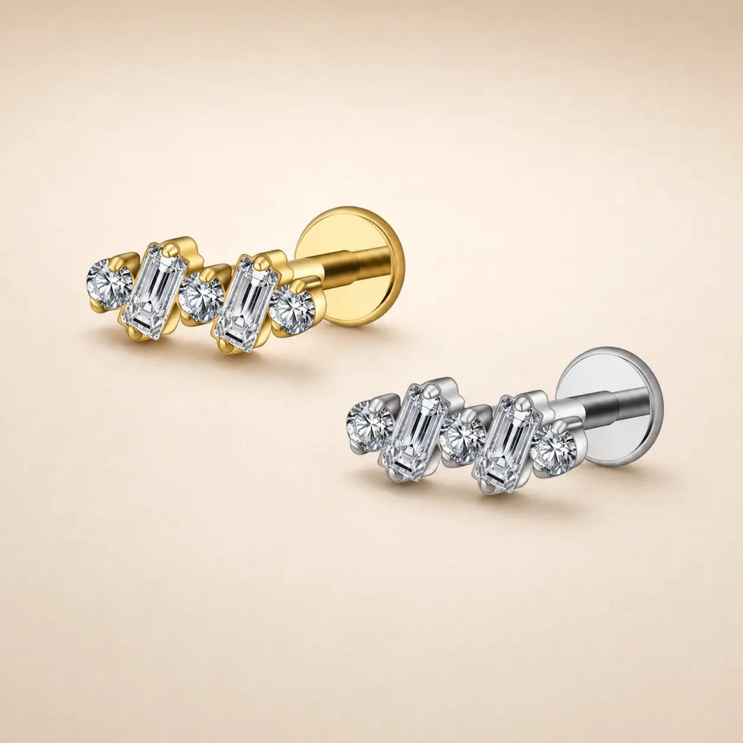 Two small stud earrings on a warm beige background, one in yellow gold and one in silver-tone metal, each featuring a row of clear baguette and round stones.
