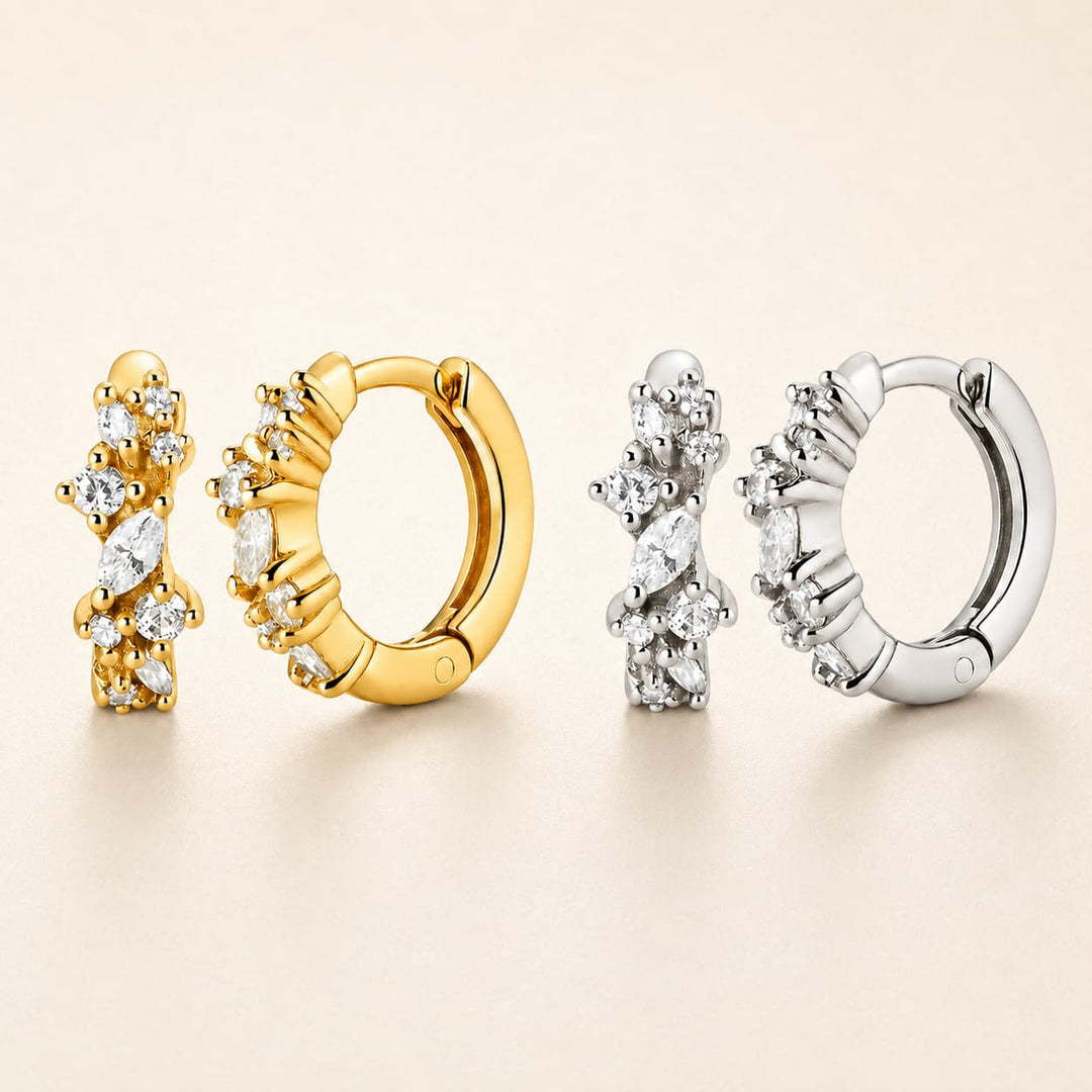 Four huggie hoop earrings arranged on a warm beige background, showing both gold and silver metal finishes with marquise-cut clear stones.
