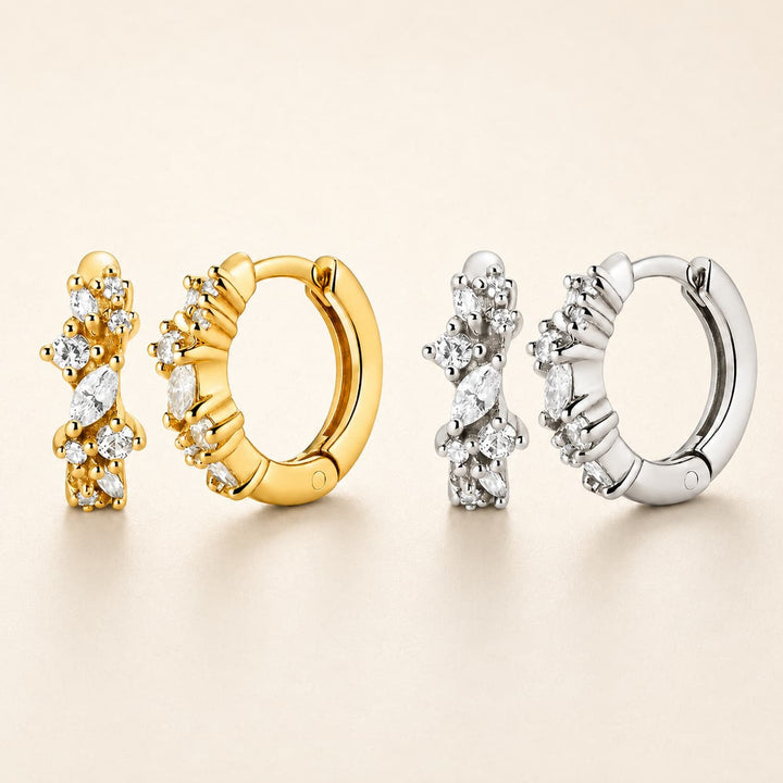 Four huggie hoop earrings arranged on a warm beige background, showing both gold and silver metal finishes with marquise-cut clear stones.
