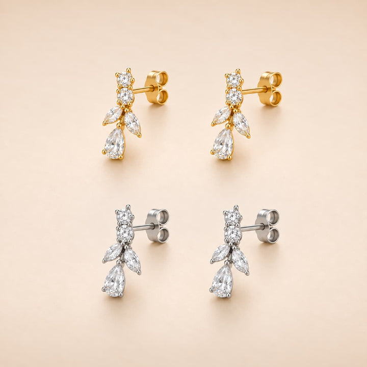 Four drop earrings arranged in a 2x2 grid on a warm beige background. The top pair features gold-toned metal with marquise-cut clear stones, and the bottom pair features silver-toned metal with marquise-cut clear stones.