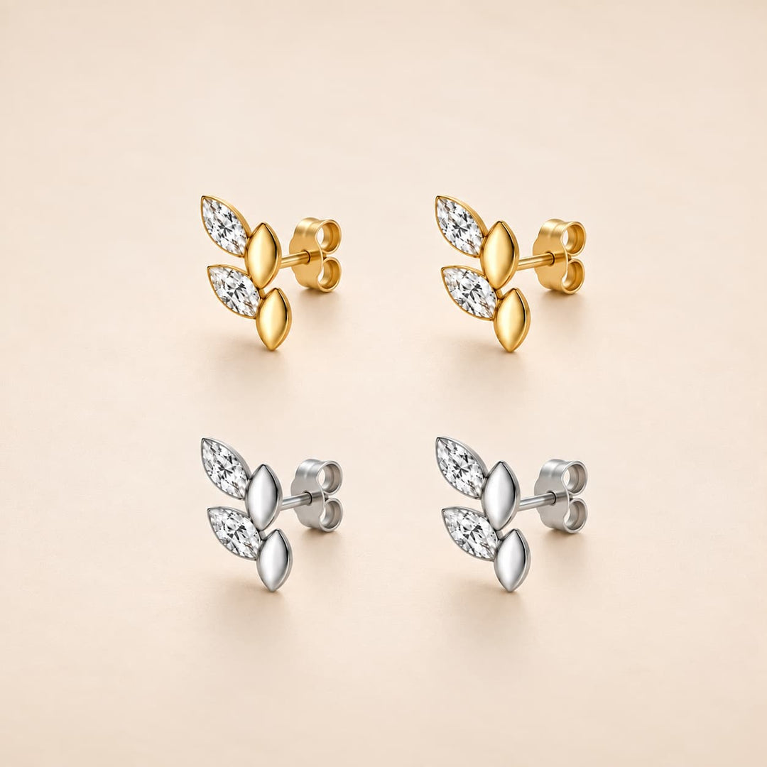 Four leaf-shaped stud earrings arranged in a 2x2 grid on a warm beige background. The top pair is yellow gold with clear marquise-cut stones, and the bottom pair is silver with matching clear stones.