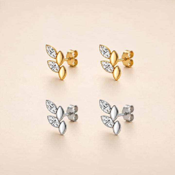 Four leaf-shaped stud earrings arranged in a 2x2 grid on a warm beige background. The top pair is yellow gold with clear marquise-cut stones, and the bottom pair is silver with matching clear stones.