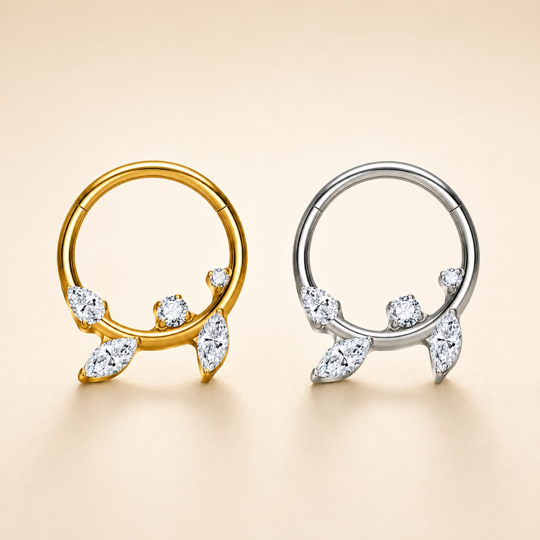 Two hoop-style body jewellery pieces with leaf-shaped crystal accents, shown side by side on a warm beige background. One is yellow gold and the other is silver, both detailed with clear marquise and round stones.