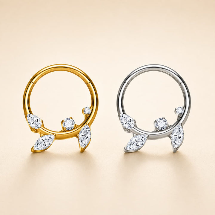 Two hoop-style body jewellery pieces with leaf-shaped crystal accents, shown side by side on a warm beige background. One is yellow gold and the other is silver, both detailed with clear marquise and round stones.