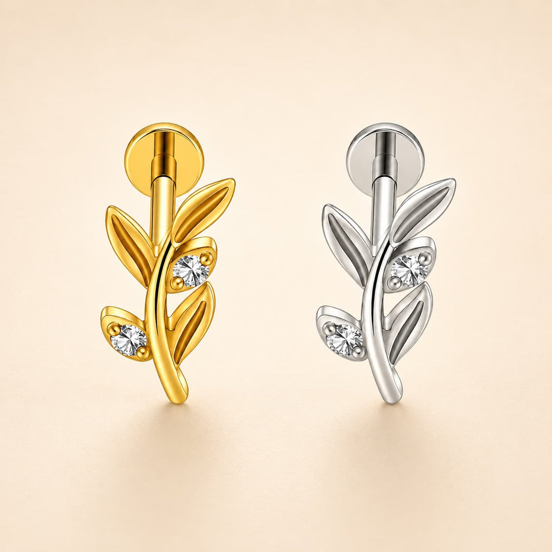 Two leaf-shaped stud earrings displayed side by side on a warm beige background. The left earring is polished yellow gold with three small clear gemstones, and the right earring is polished silver with three matching clear gemstones.