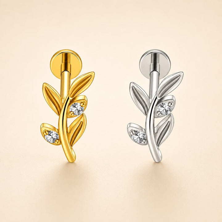 Two leaf-shaped stud earrings displayed side by side on a warm beige background. The left earring is polished yellow gold with three small clear gemstones, and the right earring is polished silver with three matching clear gemstones.