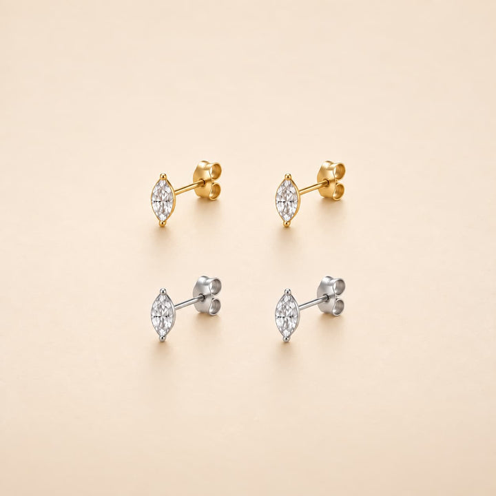 Four marquis-cut stud earrings with clear gemstones, shown in both yellow gold and silver metal finishes on a warm beige background.
