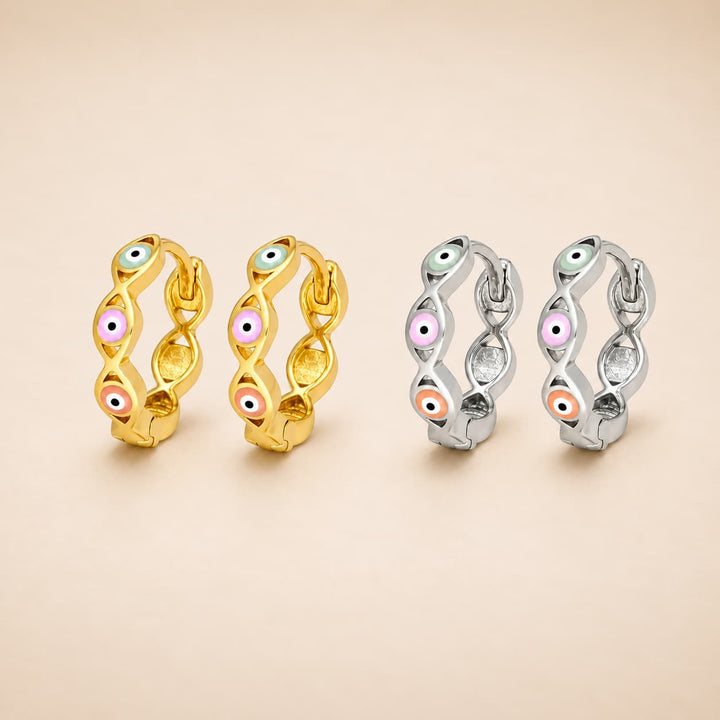 Four huggie hoop earrings with an evil eye motif, shown in two metal finishes: yellow gold on the left and silver on the right. Each earring features a twisted design with small enamel evil eye details in pink, orange, green, and white.