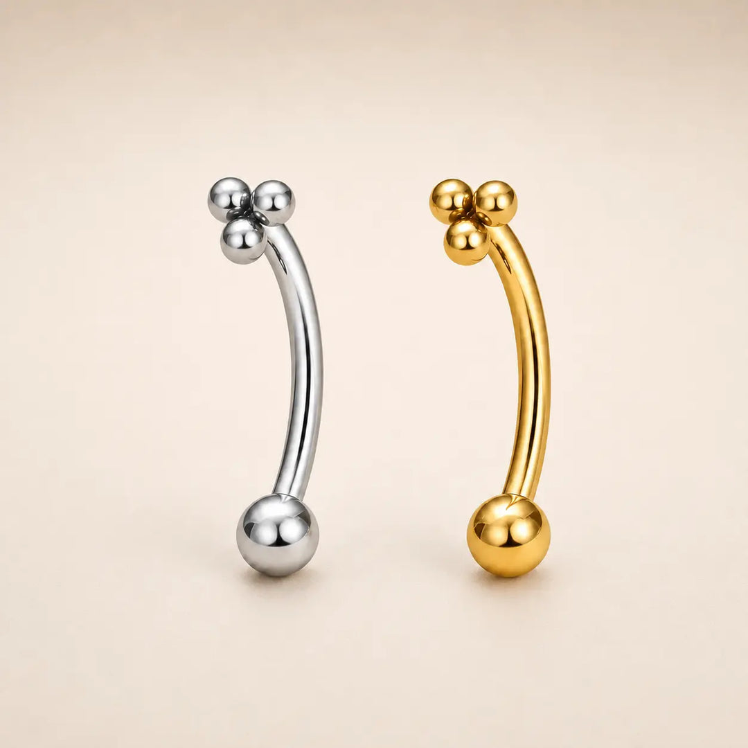 Two curved barbell-style body jewelry pieces with bead clusters at the top and a large ball at the bottom, shown on a plain warm beige background. One is silver-toned and one is gold-toned.