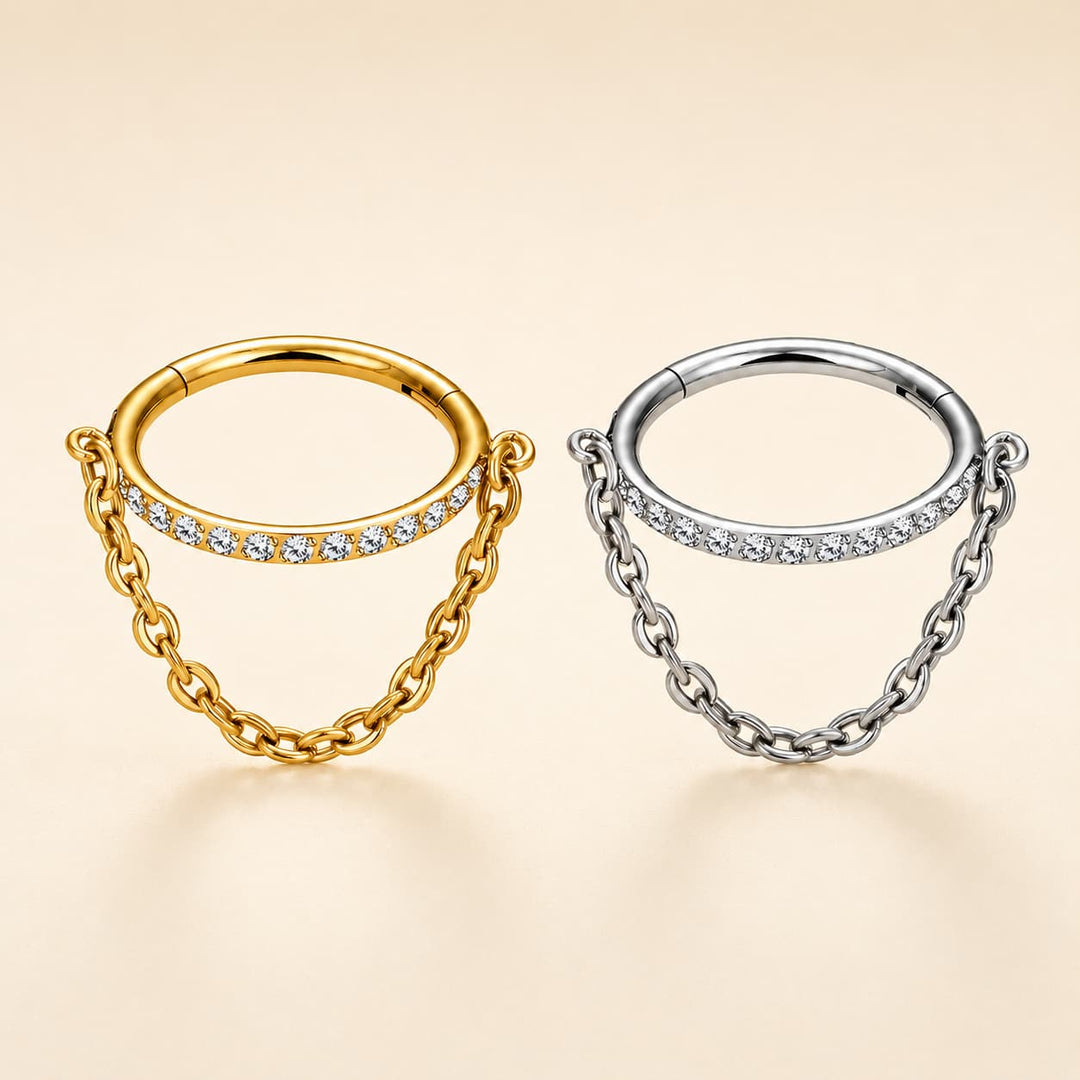 Two small hoop-style body jewelry pieces with chain accents, shown on a warm beige background. One is yellow gold with a row of clear stones, and the other is silver with a row of clear stones.