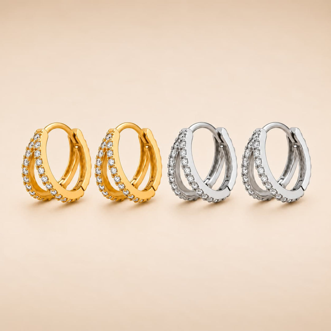 Four huggie hoop earrings arranged in two pairs, with two gold-toned hoops and two silver-toned hoops, each studded with small clear stones, on a warm beige background.