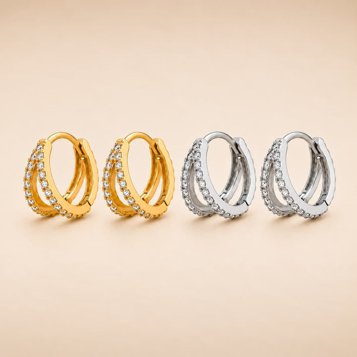 Four huggie hoop earrings arranged in two pairs, with two gold-toned hoops and two silver-toned hoops, each studded with small clear stones, on a warm beige background.