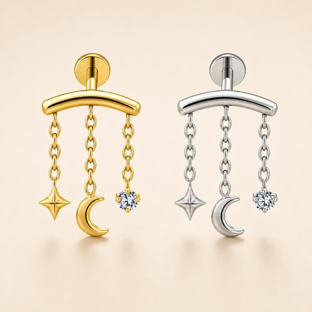Two celestial dangle earrings shown side by side on a warm beige background. The left earring is gold-toned with a curved bar, hanging chains, a crescent moon, a star, and a clear gemstone.