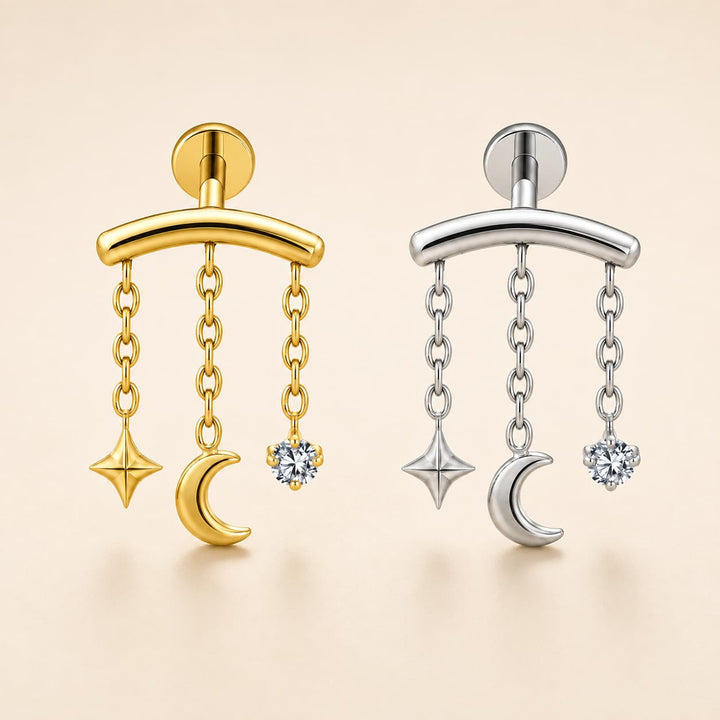 Two celestial dangle earrings shown side by side on a warm beige background. The left earring is gold-toned with a curved bar, hanging chains, a crescent moon, a star, and a clear gemstone.