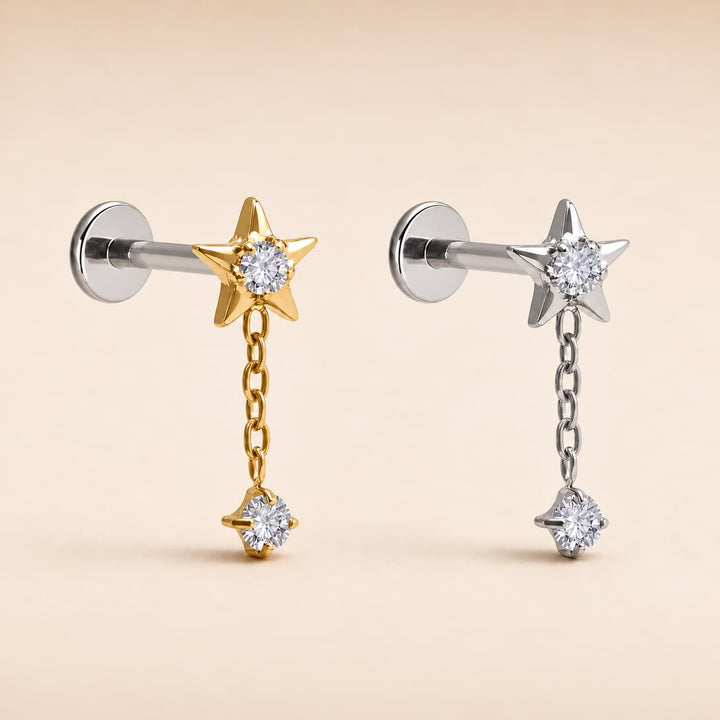 Two star-shaped dangle earrings on straight posts, one in yellow gold and one in silver, each featuring a clear round gemstone. The gold earring has a gold chain leading to a small gold star, while the silver earring has a silver chain leading to a small silver star.