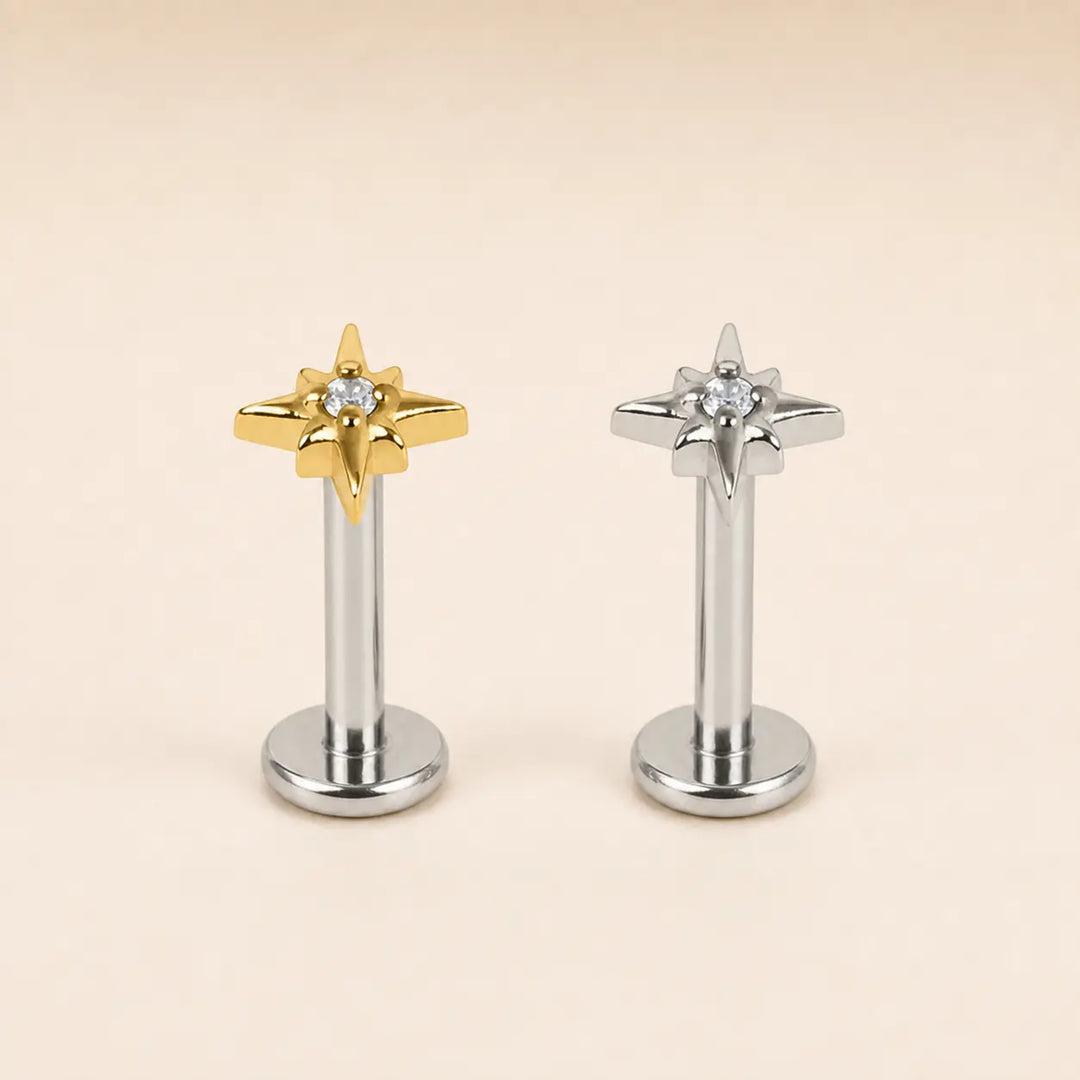 Two star-shaped stud earrings on a plain beige background, one in polished gold and one in polished silver, each with a small clear gemstone at the center.