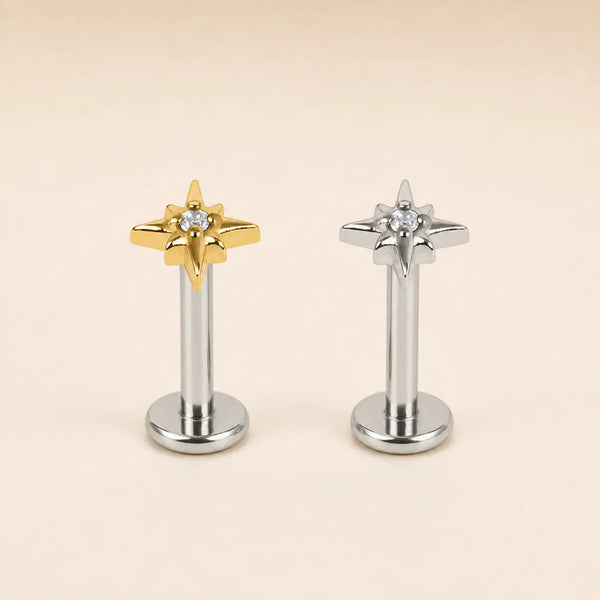 Two star-shaped stud earrings on a plain beige background, one in polished gold and one in polished silver, each with a small clear gemstone at the center.