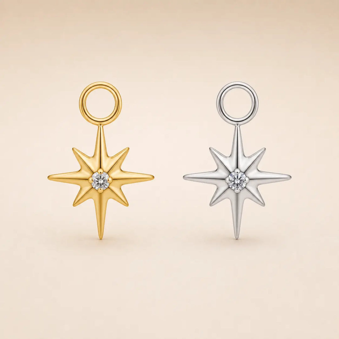 Two star-shaped pendant charms with eight-pointed rays and a small round clear gemstone at the center, shown side by side on a warm beige background. One pendant is yellow gold with a matching gold loop, and the other is silver-toned with a silver-toned loop.
