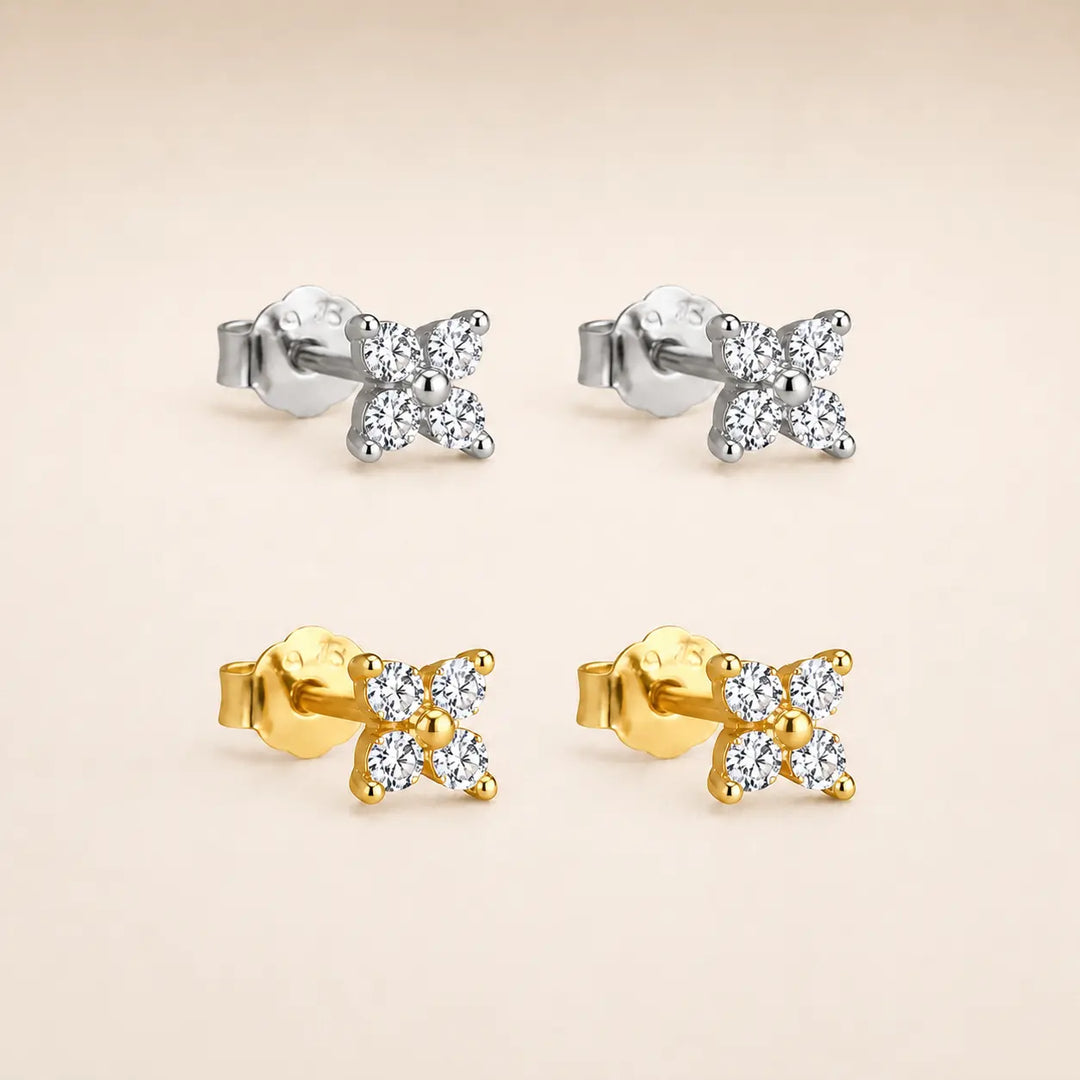 Four small bow-shaped stud earrings arranged in a 2x2 grid on a warm beige background. The top pair is silver-toned with clear stones, and the bottom pair is yellow gold-toned with clear stones.