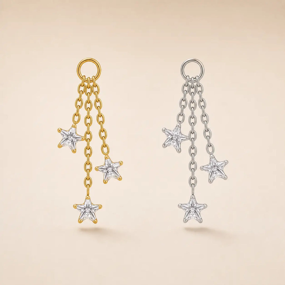 Two pairs of dangling star earrings on a warm beige background. One pair is gold-toned with three dangling star charms, and the other is silver-toned with three matching star charms.