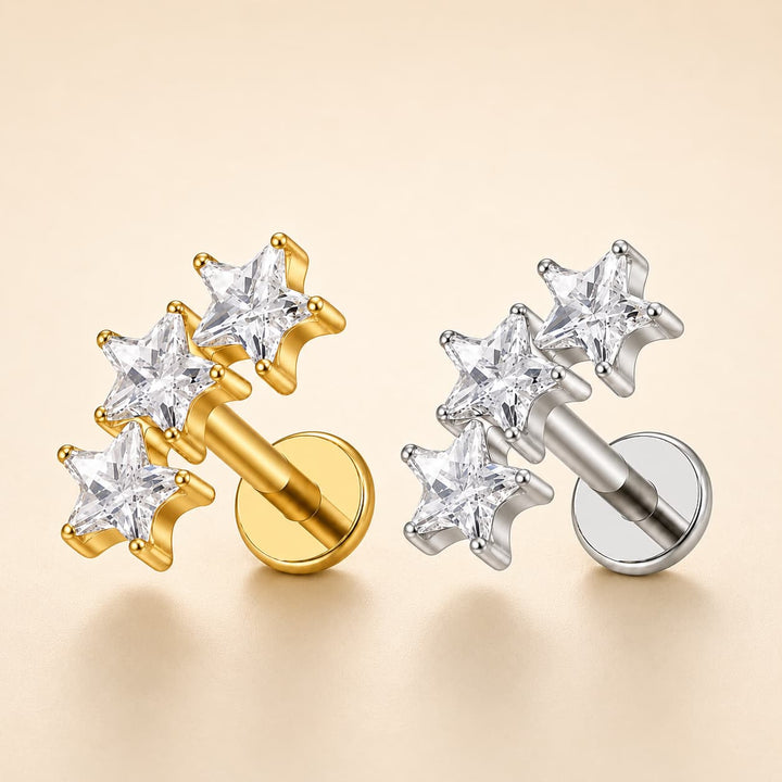 Three star-shaped stud earrings shown on a warm beige background, with one gold-tone pair and two silver-tone pairs. Each earring features three clear gemstone stars set in a polished metal post with a round backing.